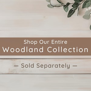 May include: A rectangular brown banner with white text that reads Shop Our Entire Woodland Collection and Sold Separately, positioned over a light-colored wooden surface with a sprig of green eucalyptus leaves in the upper right corner.