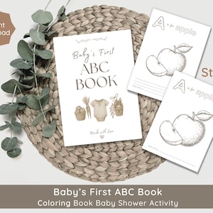 May include: A baby shower activity set is displayed. It features a "Baby's First ABC Book" and two "A is for apple" colouring pages. The items are arranged on a woven mat with eucalyptus leaves. The words "Instant Download" are visible.