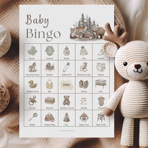 A crocheted deer toy with brown antlers and a white and beige body sits next to a "Baby Bingo" game card. The card features baby-themed illustrations and the words "Baby Bingo" in a soft, neutral font. Two yarn balls are also in the image.