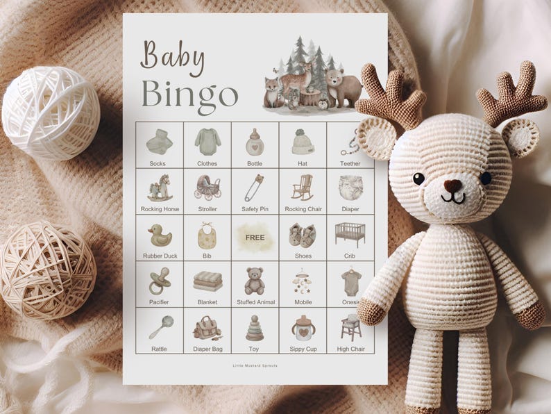 A crocheted deer toy with brown antlers and a white and beige body sits next to a "Baby Bingo" game card. The card features baby-themed illustrations and the words "Baby Bingo" in a soft, neutral font. Two yarn balls are also in the image.