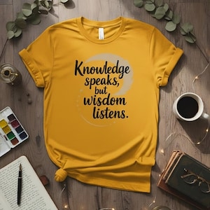 Peut inclure: T-shirt jaune moutarde avec la citation "Knowledge speaks, but wisdom listens" en écriture noire. Le t-shirt est présenté sur une surface en bois avec une tasse de café, des livres et du matériel d'art.