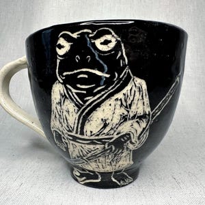 May include: Black ceramic mug with a white frog illustration wearing a kimono and holding a sword. The mug has a white handle and a wide, rounded shape. The frog design is detailed and artistic.