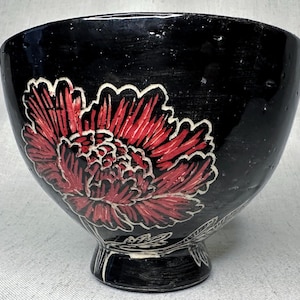 May include: A black ceramic bowl with a red and white floral design. The bowl has a wide, rounded shape with a pedestal base. The flower is a large peony, with detailed petals and a white outline. The bowl is likely handmade and has a rustic aesthetic.