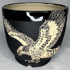 May include: A black ceramic planter featuring a cream-colored sgraffito illustration of an eagle in flight. The exterior has a matte black finish with a glossy black rim, while the interior is a smooth, light cream color with a circular base.