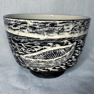 May include: A black and white ceramic bowl with a boat and water design. The bowl features a detailed illustration of a boat sailing on wavy water. The rim and base are black, while the interior is white.