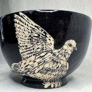 May include: A glossy black ceramic bowl featuring a cream-colored sgraffito illustration of a bird with its wings spread wide. The bird design covers the front of the bowl, showcasing detailed feather textures and a distinct eye, set against the dark finish.