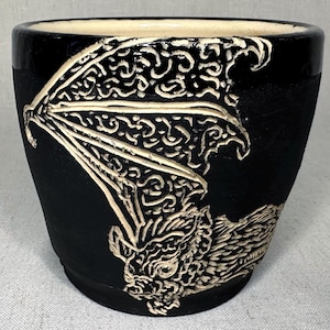 May include: A black ceramic planter featuring a carved, cream-colored bat design on the exterior. The bat has detailed wings and a textured face. The interior of the pot is a smooth, light cream color, contrasting with the matte black exterior finish.