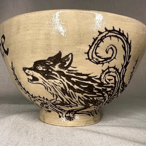 May include: Cream-colored ceramic bowl with a black wolf illustration and thorny vine design. The bowl has a wide opening and a small, rounded base. The wolf is depicted with an open mouth, and the vines curl around the bowl's exterior.