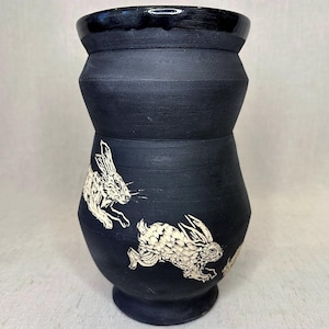 May include: A matte black ceramic vase with a wide, flared rim and a tapered base. The exterior surface features two white, etched illustrations of rabbits in motion, showcasing detailed fur textures and dynamic poses against the dark clay body.