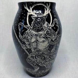 May include: Black ceramic vase with a detailed white illustration of a deer-like figure with antlers, a halo, and holding a sword. The vase has a glossy finish and a rounded shape, suitable for home decor.
