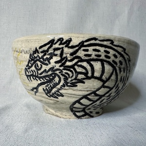 May include: A ceramic bowl with a cream-colored base and a black dragon design. The dragon is hand-painted and wraps around the bowl's exterior. The bowl has a wide opening and a rounded base.