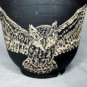 May include: A matte black ceramic vessel featuring a carved, cream-colored owl with its wings fully extended across the front. The owl design displays intricate feather detailing and a textured surface, contrasting sharply against the dark background.