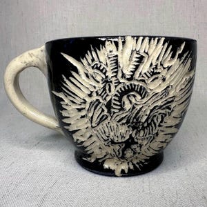 May include: A black ceramic mug with a cream-colored handle and a detailed, raised white design. The design appears to be an abstract, stylized image. The mug is a unique piece of drinkware.