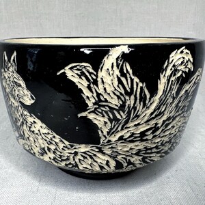 May include: A black ceramic bowl featuring a carved white relief design of a multi-tailed fox. The fox is depicted in profile with textured fur details and several large, sweeping tails that wrap around the side of the rounded vessel.