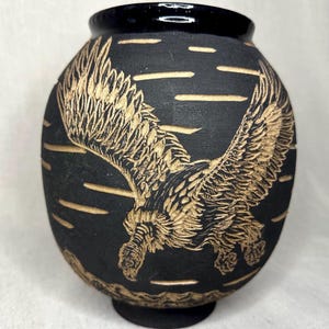 May include: A matte black ceramic vase featuring a hand-carved design of a large eagle in flight with its wings spread wide. The intricate engraving reveals a lighter tan color beneath the dark surface, depicting the bird and horizontal lines.