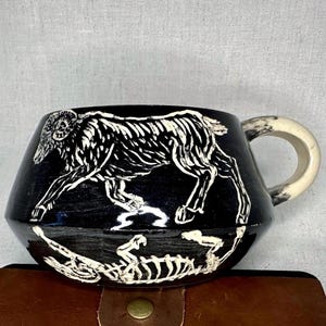 May include: A black ceramic mug featuring a white graphic illustration of a ram in profile above a skeletal depiction of the same animal. The mug has a wide, tapered body and a curved, off-white handle with dark, mottled markings on its surface.