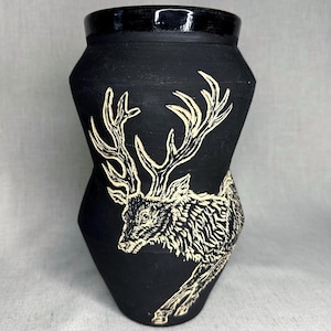 May include: A matte black ceramic vase with a wide, angular midsection and a glossy black rim. The exterior features a detailed, cream-colored sgraffito illustration of a deer with large, branching antlers, covering the front of the vessel.