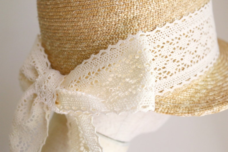 A straw hat with a wide brim and a white lace trim. The lace is tied in a bow at the back of the hat.