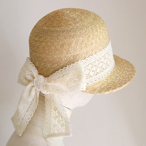 A straw hat with a wide brim and a white lace ribbon tied in a bow. The hat is beige and the ribbon is white. Made by Grosgrain