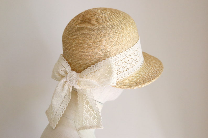 A straw hat with a wide brim and a white lace ribbon tied in a bow. The hat is beige and the ribbon is white. Made by Grosgrain