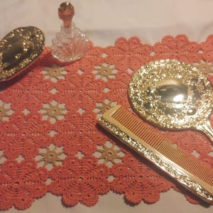 May include: Three gold-toned hand mirrors with ornate designs, one with a comb, on a pink crocheted doily.
