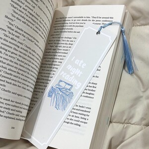 Op de afbeelding: Een wit boekenlegger met de tekst "I ate night reading" en een blauwe illustratie van een bed. De boekenlegger zit in een boek met tekst en een blauwe kwast.