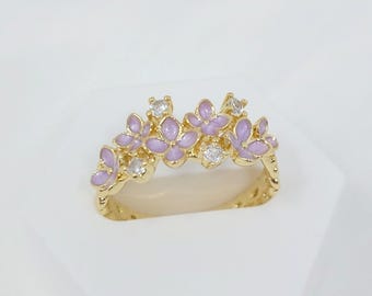 May include: A gold-toned ring featuring a floral design with lavender enamel petals and clear, sparkling gemstones. The ring is set on a white surface, showcasing its intricate details and elegant style.