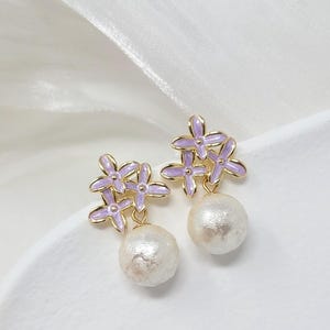 May include: A pair of gold-toned earrings featuring clusters of lavender-colored flower-shaped accents. Each earring has a large, round, off-white pearl-like bead dangling below the floral design. The earrings are set against a white background.
