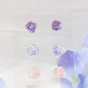 May include: A clear acrylic display features six small rose-shaped earrings. The earrings are in three pairs, each pair in a different color: purple, iridescent, and pink. The roses have gold-colored accents and are set against a soft, blurred background.