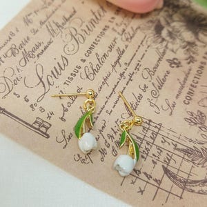 May include: Gold-coloured earrings with white lily of the valley flowers and green leaves. They are set against a vintage paper background with French text and a key illustration.