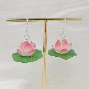 Peut inclure: Une paire de boucles d'oreilles en forme de fleur de lotus. Chaque boucle d'oreille présente une fleur de lotus rose et blanche avec une base de feuille verte. Les boucles d'oreilles sont présentées sur un support doré sur fond blanc.