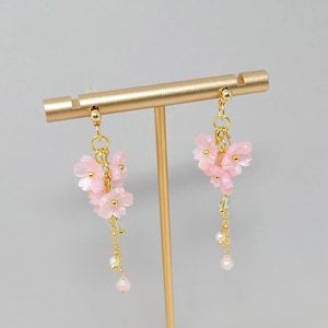 May include: Dangle earrings with pink flower charms and gold-coloured metal detailing. The earrings have small pearl and green bead dangles. They are displayed on a gold-coloured earring stand. The earrings are a delicate accessory.