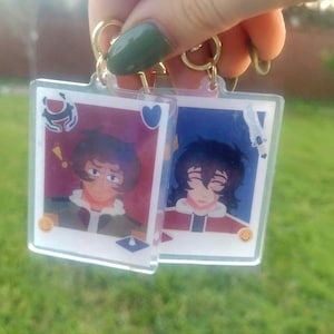 May include: Two clear acrylic keychains with gold-colored rings. Each keychain features a cartoon illustration of a person with brown hair and a red and white outfit. The background of each keychain is a different color.