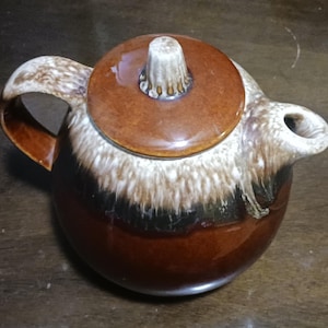 May include: A brown and white glazed ceramic teapot with a lid. The teapot has a brown handle and a brown spout.