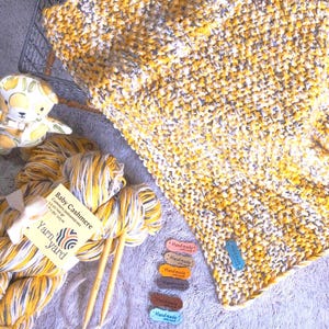 May include: A hand-knitted baby blanket in yellow, grey, and white, with a matching toy and yarn. The yarn is labelled "Baby Cashmere" and includes knitting needles and colour tags. A wire basket is also in the image.