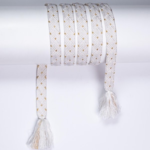 May include: A long, woven friendship bracelet featuring a repeating diamond pattern in white and metallic gold thread. The bracelet is wrapped multiple times around a cylindrical object and terminates at both ends with white and gold fringe tassels.