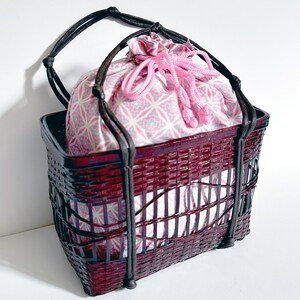 May include: A burgundy woven basket with a black handle and a pink patterned drawstring bag inside. The bag is tied with a pink cord. The basket is made of woven material.