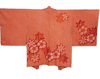 May include: A coral-colored haori jacket featuring a textured fabric surface and an embroidered floral pattern in shades of pink and red. The garment has wide, rectangular sleeves and a central opening, displaying a repeating cherry blossom motif.