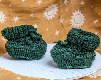 May include: A pair of dark green crocheted baby booties with a fold-over cuff. The booties are sitting on a white wooden surface.