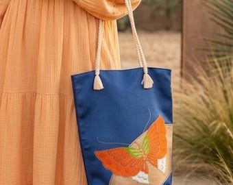 May include: A long-sleeved, tiered peach-colored maxi dress with subtle embroidery on the bodice and sleeves, paired with a navy blue tote bag featuring a large orange and green butterfly graphic and thick white rope handles.