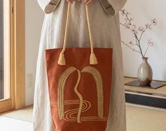 May include: A long-sleeved, floor-length beige linen dress with a round neckline, paired with a rust-colored tote bag featuring yellow rope handles and a yellow embroidered arch design with wavy lines on the front surface of the bag.