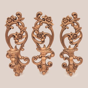 May include: A set of three matching wall sconces in a metallic bronze finish. Each sconce features an elongated vertical frame with intricate scrollwork, carved floral details at the top and bottom, and a central circular candle holder cup.