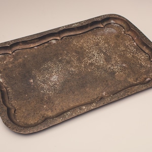 May include: A rectangular metal serving tray with a dark, tarnished finish and a scalloped edge. The surface features an intricate, embossed floral pattern that covers the central area and extends along the raised, decorative border.