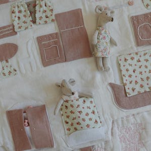 Puede incluir: Un tapete de juego hecho a mano con un dise&ntilde;o caprichoso. Presenta varias aplicaciones de tela en tonos rosa y blanco, incluyendo cortinas en miniatura, una mesa y un barco. Tambi&eacute;n hay dos peque&ntilde;as mu&ntilde;ecas de rat&oacute;n.