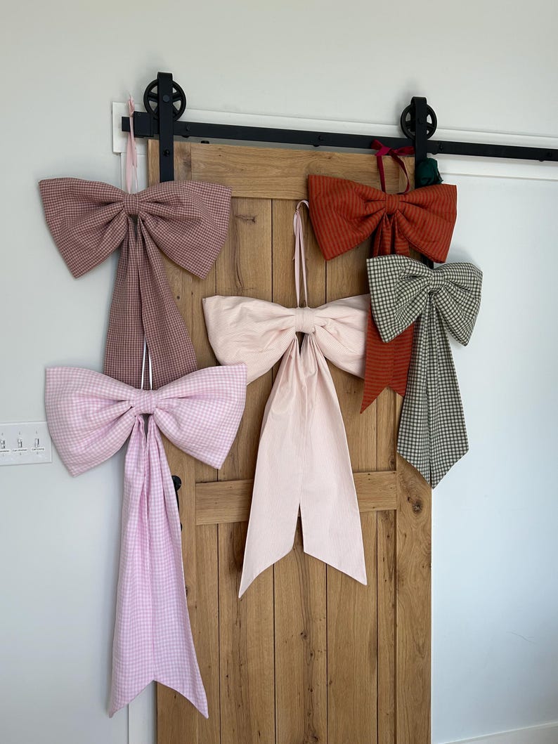 K&ouml;nnte beinhalten: F&uuml;nf verschiedene Gingham-Schleifen in verschiedenen Farben, die an einer Holzt&uuml;r h&auml;ngen. Die Schleifen sind aus Stoff und haben ein kariertes Muster. Die Schleifen sind alle auf die gleiche Weise gebunden, wobei die Schleifenenden nach unten h&auml;ngen.