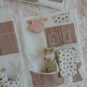 Puede incluir: Un tapete de juego hecho a mano de tela blanca, con detalles rosas y florales. Incluye una ba&ntilde;era, una lavadora y una cama, con un peque&ntilde;o juguete de rat&oacute;n en la ba&ntilde;era. El tapete est&aacute; decorado con tela rosa y floral.