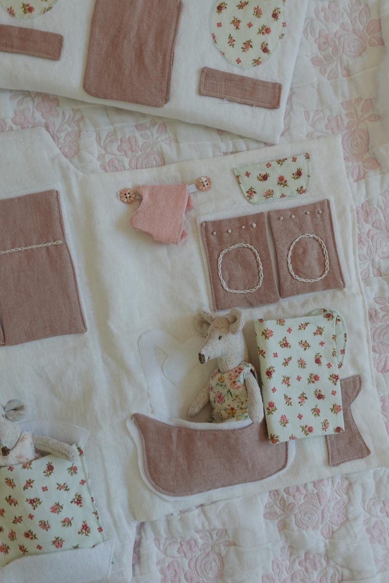 Puede incluir: Un tapete de juego hecho a mano de tela blanca, con detalles rosas y florales. Incluye una ba&ntilde;era, una lavadora y una cama, con un peque&ntilde;o juguete de rat&oacute;n en la ba&ntilde;era. El tapete est&aacute; decorado con tela rosa y floral.