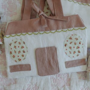 Puede incluir: Un bolso de mano de tela blanca y rosa hecho a mano con forma de casa, con detalles florales y un ribete festoneado verde. El bolso tiene una puerta y dos ventanas ovaladas con tela floral. Dos peque&ntilde;as mu&ntilde;ecas rat&oacute;n con vestidos florales est&aacute;n al lado del bolso.