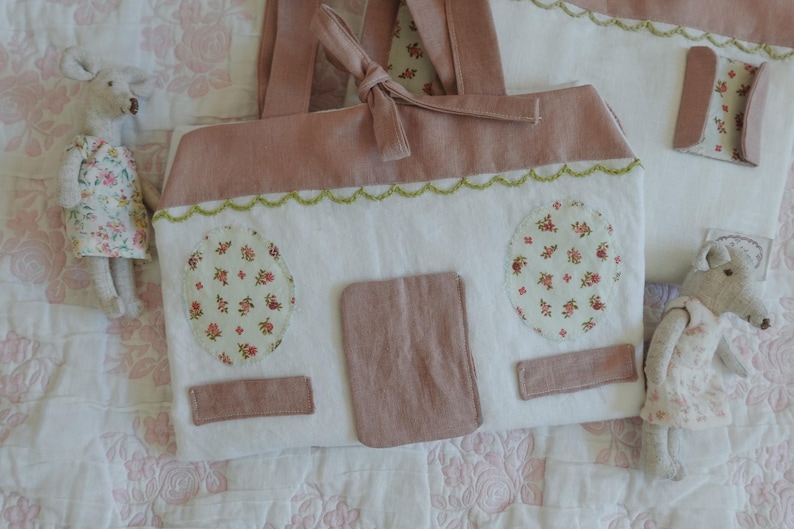 Puede incluir: Un bolso de mano de tela blanca y rosa hecho a mano con forma de casa, con detalles florales y un ribete festoneado verde. El bolso tiene una puerta y dos ventanas ovaladas con tela floral. Dos peque&ntilde;as mu&ntilde;ecas rat&oacute;n con vestidos florales est&aacute;n al lado del bolso.