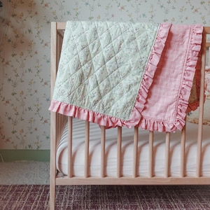 Pode incluir: Uma colcha de bebê verde claro e rosa com um padrão floral e acabamento de babados rosa sobre um berço de madeira. A colcha tem um design acolchoado e um padrão xadrez rosa no verso.
